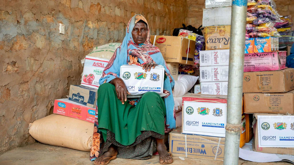 Woman in front of her own shop. The shop is one room with banana cases full of things to sell.